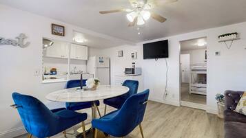 A modern kitchen with white cabinets, a microwave, and a sink. There is a dining area with a round table and blue chairs. A ceiling fan is visible above the dining area.