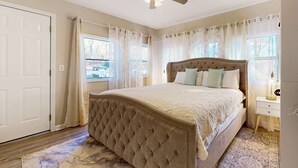 Ein Schlafzimmer mit einem gepolsterten Kopfteil, einem großen Bett, einem Deckenventilator und Blick ins Freie durch ein Fenster mit durchsichtigen Vorhängen.