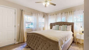 A bedroom with a tufted headboard, a large bed, a ceiling fan, and a view of the outside through a window with sheer curtains.