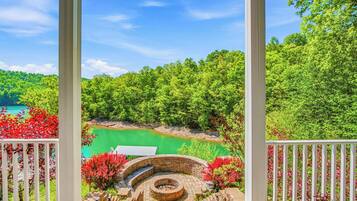 A gazebo with a circular brick design, surrounded by red flowering plants and a view of a green lake.
