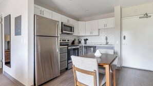 A modern kitchen with stainless steel appliances, white cabinetry, and a dining area with a wooden table and chair. - Landing | Spectacular Studio, Gym, Yoga Studio (Port Charlotte)