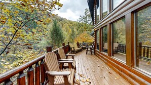 A wooden deck with Adirondack chairs, a table, and a view of a forested hillside. - Upscale Cabin W Hot Tub, Sleeps 22・Game Room・Pool (Sevierville)