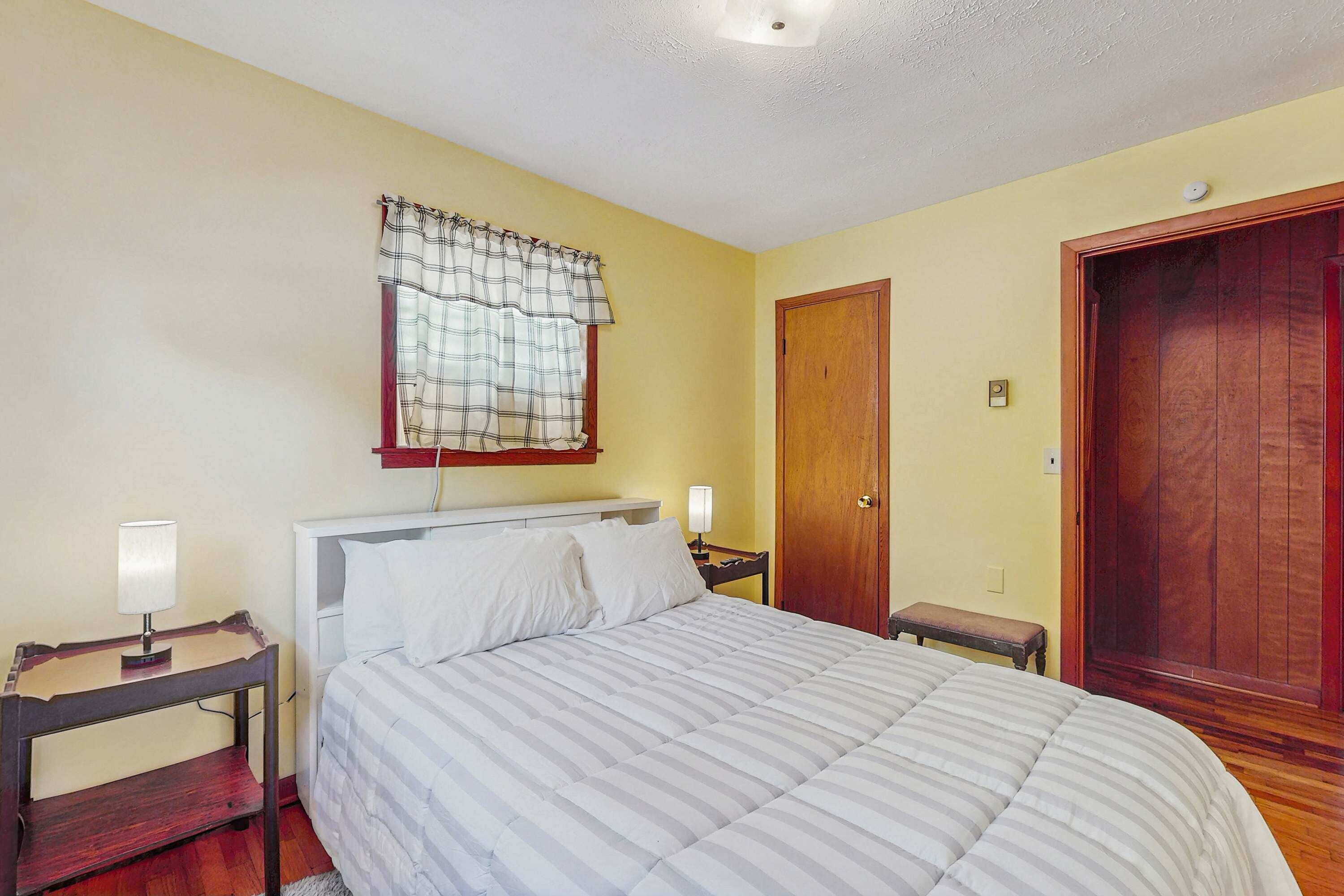 Un dormitorio con una cama, mesitas de noche, un armario auxiliar, una puerta de madera y una ventana con cortinas.