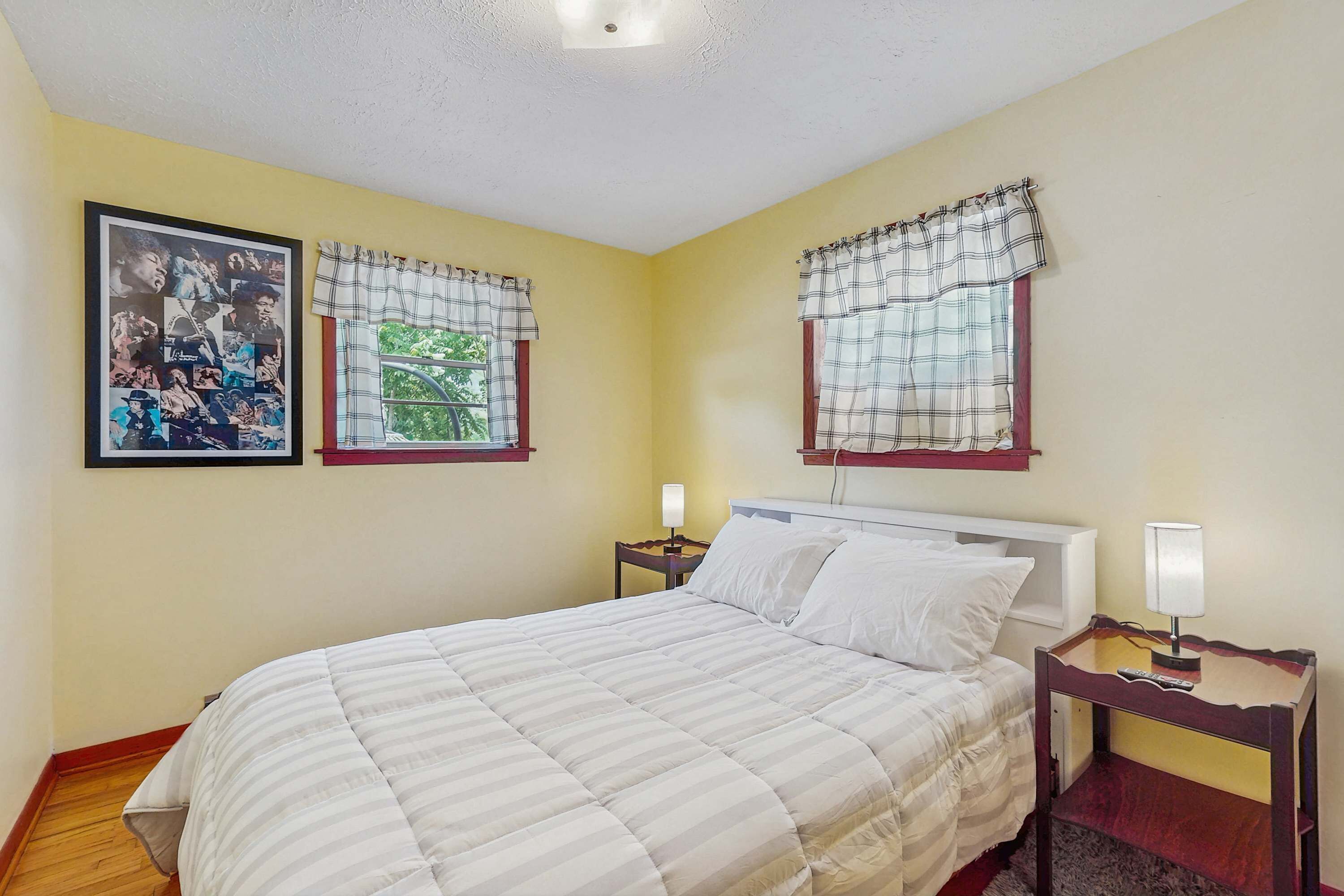 Una cama bien hecha en una habitación con una pared amarilla, un collage enmarcado a la izquierda y una ventana con cortinas escocesas.