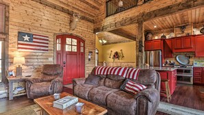 Un interno rustico con soffitto in legno, una porta rossa, un divano in pelle, un tavolino da caffè in legno e un angolo cucina con mobili rossi ed elettrodomestici in acciaio inossidabile.