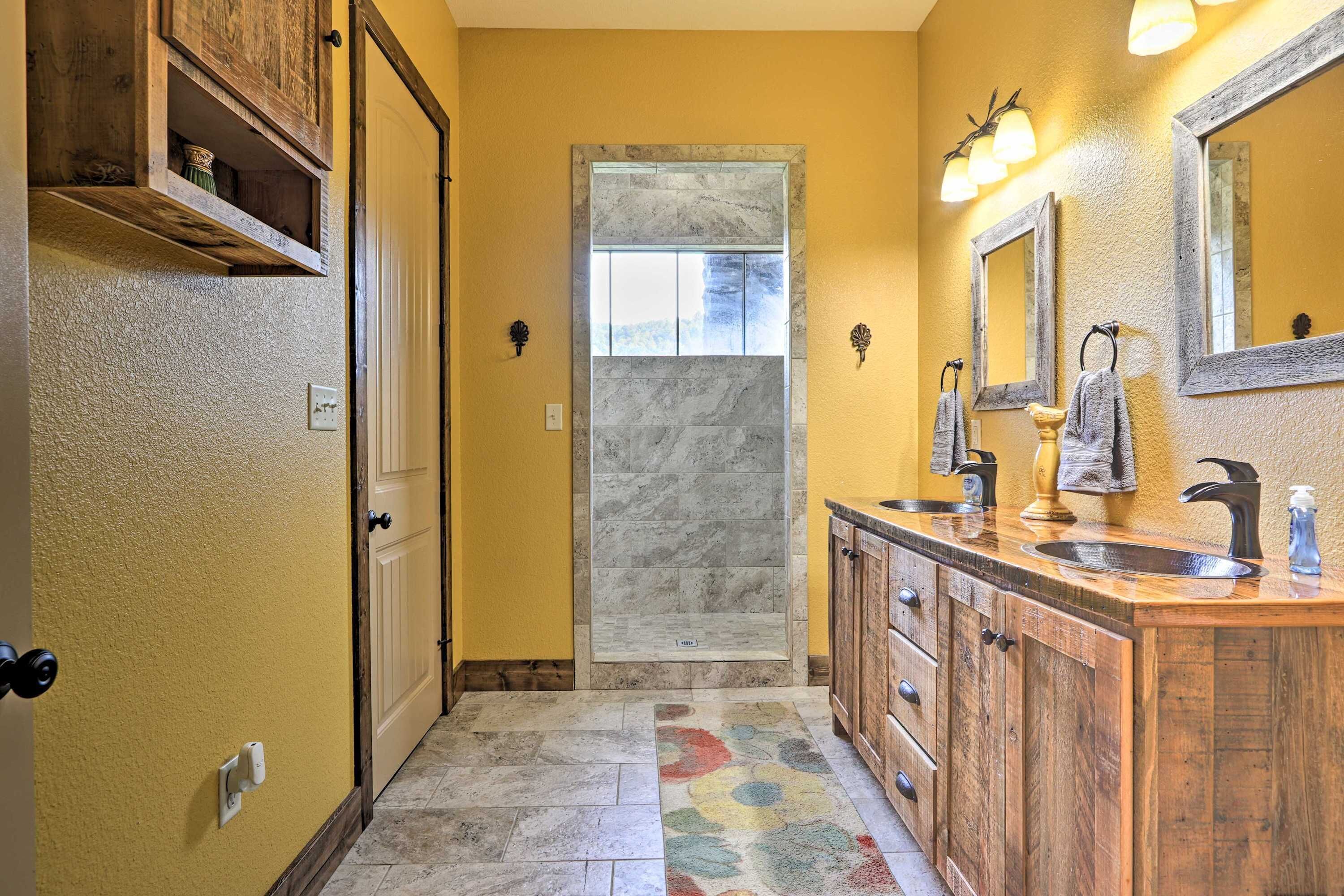 A bathroom with a wooden vanity, two sinks, a mirror, and a shower with a marble wall.