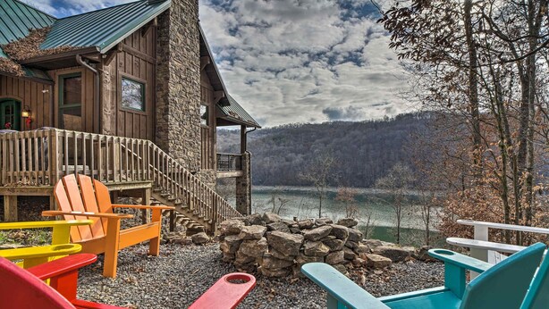 Una baita in legno con rivestimento in pietra, un pontile di legno con poltrone Adirondack colorate, e vista su un lago e una foresta.