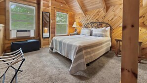 Une chambre dans une cabane en bois, avec un lit, deux fenêtres équipées de stores, un ventilateur de plafond et une petite table avec une lampe.