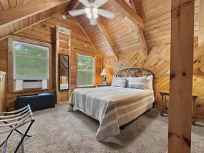 Ein Schlafzimmer einer Holzhütte mit einem Bett, zwei Fenstern mit Jalousien, einem Deckenventilator und einem kleinen Tisch mit einer Lampe.