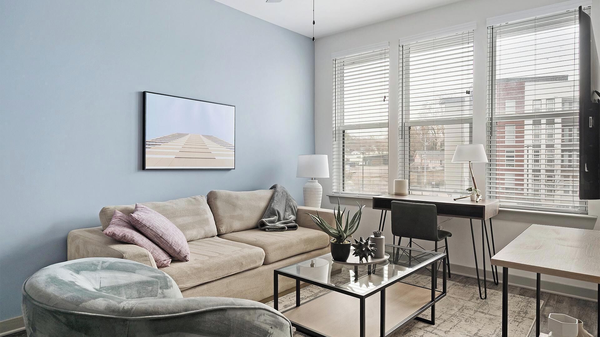 A modern living room with a sofa, a glass coffee table, a chair, and a desk with a lamp. There is a window with blinds, a framed picture on the wall, and a potted plant on the table.