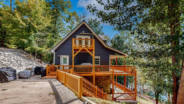A cabin with a wooden deck, a stone foundation, and a view of the surrounding forest. - Lakefront Cabin, Sleeps 14・2 Level Dock・Sun Deck (LaFollette)