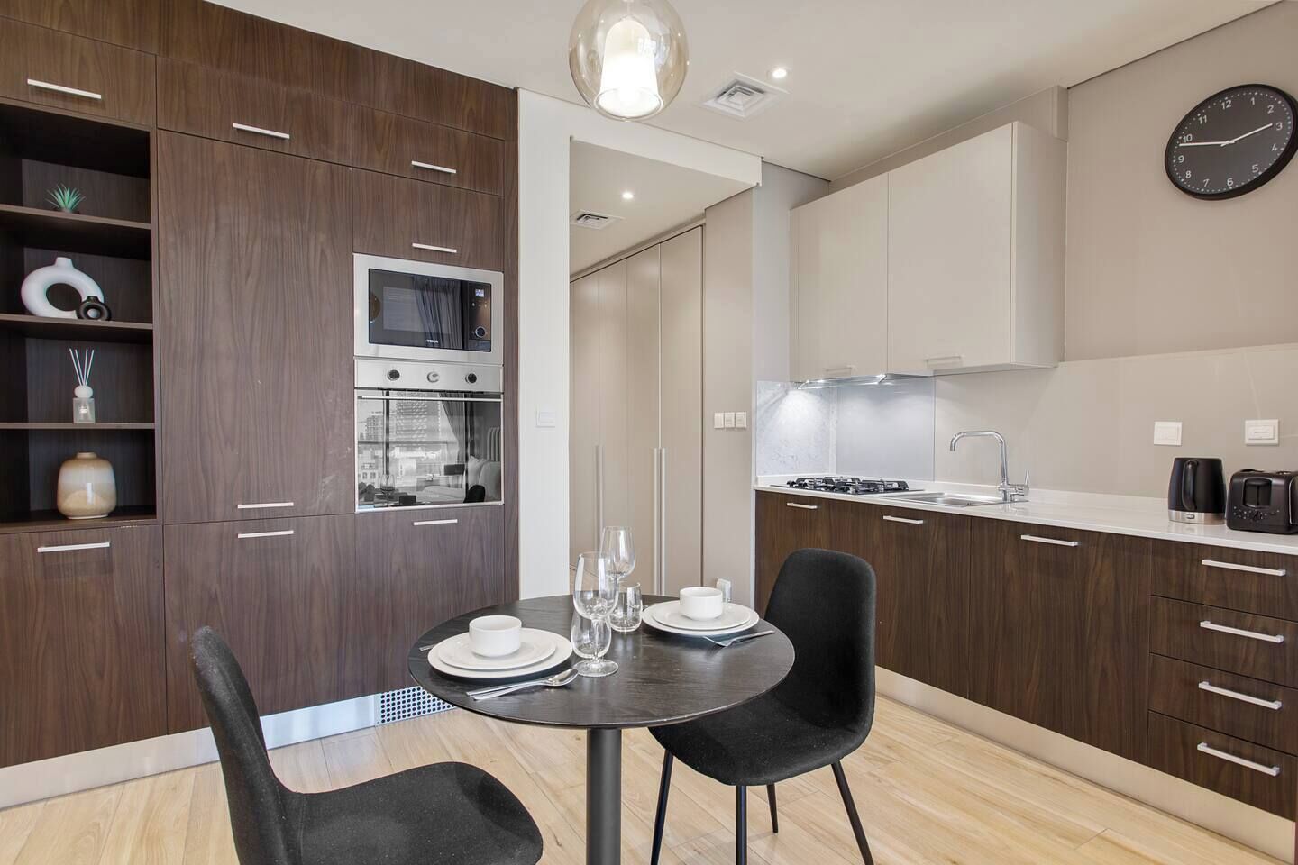 Une cuisine moderne avec des meubles de couleur bois foncé, un four encastré et un coin repas comprenant une table ronde et deux chaises.