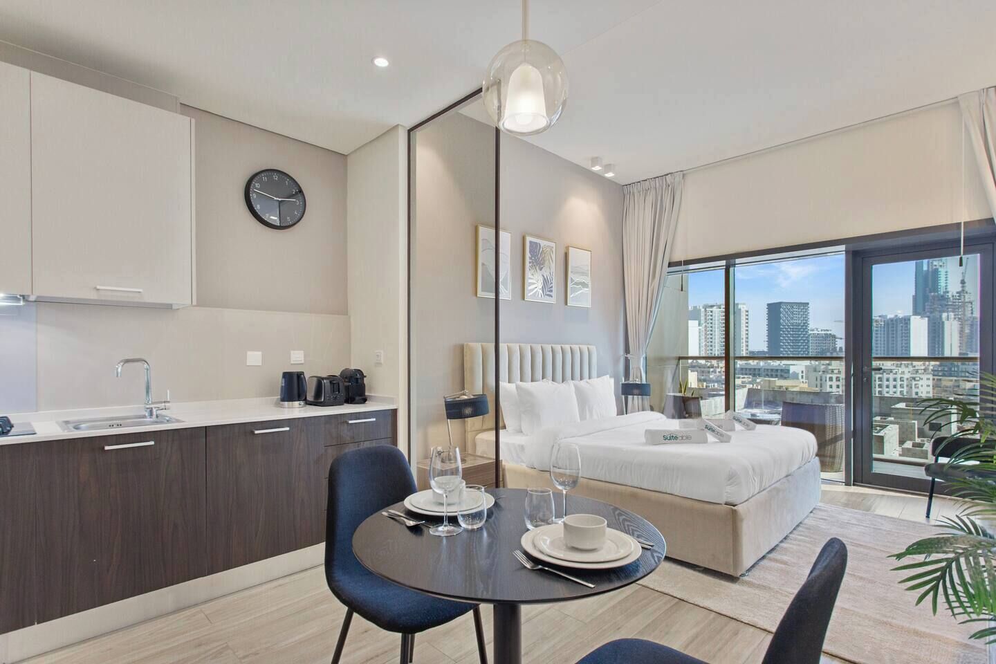 A modern hotel room with a kitchenette, a dining area, a bed with a city view, and a large window.