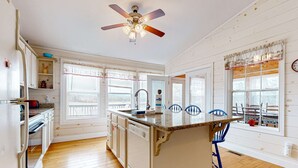 Une cuisine avec un plancher en bois, des armoires blanches, un îlot central, des chaises bleues et un ventilateur de plafond.