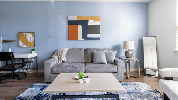 En moderne stue med en grå sofa, et sofabord, et skrivebord med stol, en lampe og et stort abstrakt billede på væggen.