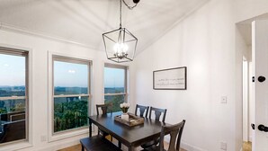 Un coin repas avec une table en bois, six chaises et un lustre. De grandes fenêtres offrent une vue sur la verdure et le paysage urbain.