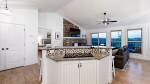 Une cuisine spacieuse dotée d’un grand îlot central, de meubles blancs et d’un mur d’accent en pierre. On y trouve également un ventilateur de plafond et de grandes fenêtres qui offrent une vue sur l’extérieur.