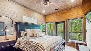 Un dormitorio con cama, mesitas de noche, ventilador de techo y ventanas amplias con vistas a árboles.