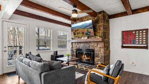 Ein gemütliches Wohnzimmer mit Kamin aus Stein, einem grauen Sofa, einem Holzstuhl und einem großen an der Wand befestigten Fernseher.
