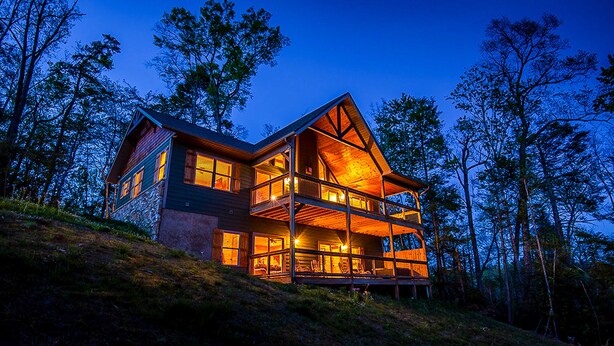 Une grande cabane en rondins à plusieurs étages, dotée d’un balcon imposant et de grandes fenêtres, et entourée d’arbres.