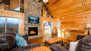 L’intérieur chaleureux d’un chalet avec une cheminée en pierre, des murs en bois, un téléviseur fixé au-dessus de la cheminée et un coin salon confortable avec un canapé et un fauteuil.