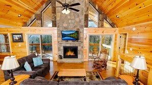 L’intérieur chaleureux d’un chalet avec une cheminée en pierre, des meubles en bois et de grandes fenêtres offrant une vue panoramique.