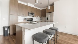 A modern kitchen with wooden cabinets, stainless steel appliances, a white countertop, and three grey bar stools. - Landing | Stunning 2BD, Pool, Clubhouse (Miromar Lakes)
