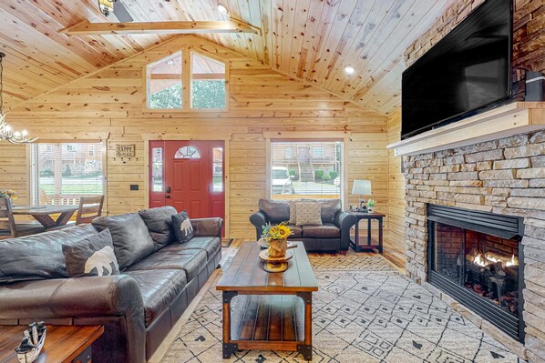 A cozy log cabin interior with a leather sofa, a stone fireplace, and a television mounted on the wall. - Serene & Spacious Log Cabin・Hot Tub・Screened Porch (Gatlinburg)