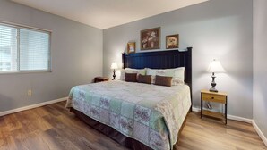 Une chambre à coucher comprenant un lit, deux tables de chevet, une commode, une fenêtre avec des stores et des œuvres d’art accrochées au mur.