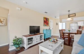 Un salon avec un téléviseur, une table basse, un coin repas avec des chaises et, au fond, une cuisine.