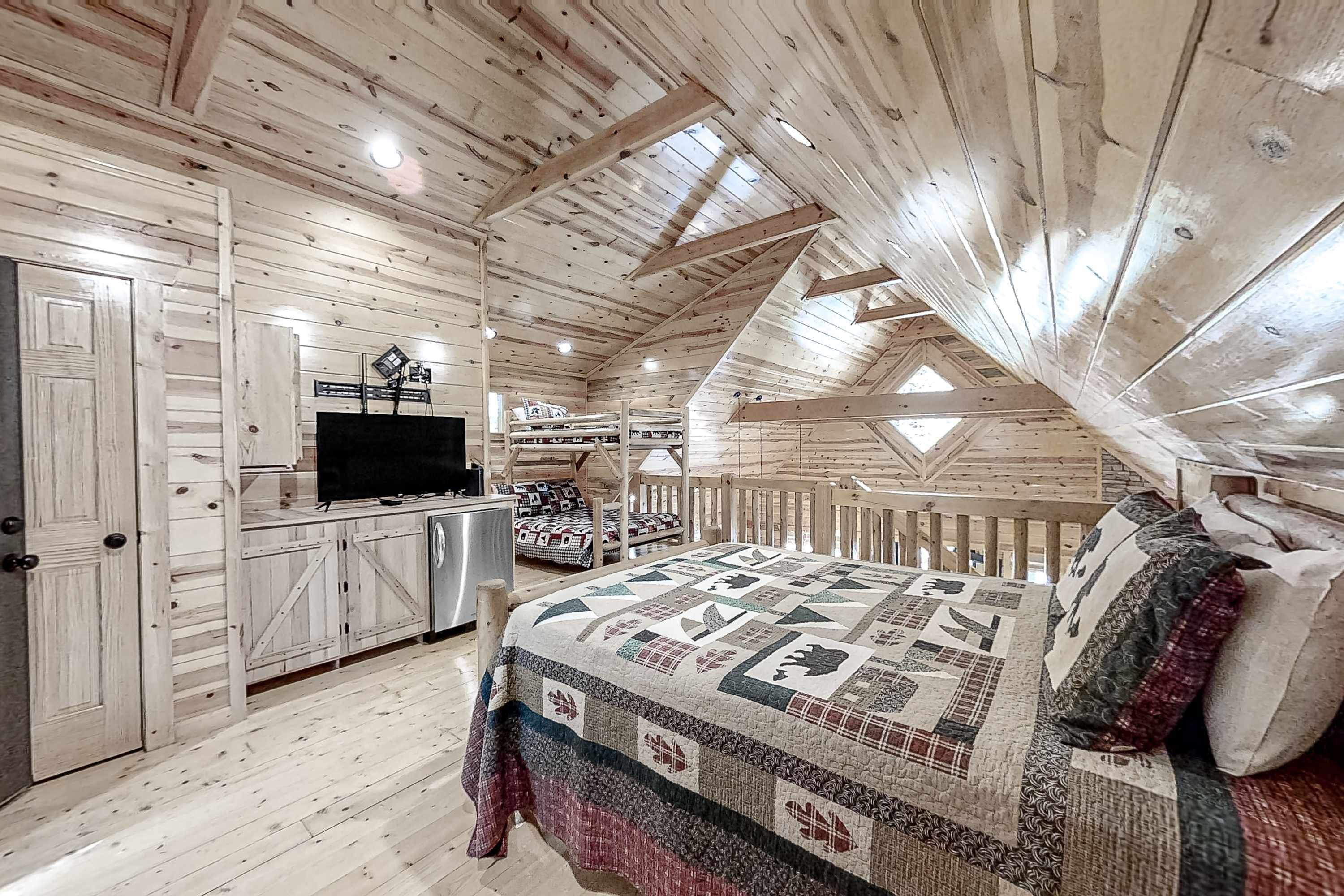 A cozy attic bedroom with a wooden ceiling, a bed with a quilt, a desk with a computer, and a TV.