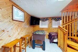 Ein gemütliches Zimmer mit Holzwänden, einem Kicker, einem Bett mit lila Decke und einer Holztreppe.