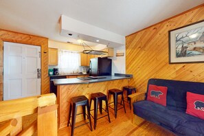 Eine kompakte Küche mit einer zentralen Kochinsel, Holzwänden und einem dunkelblauen Sofa mit Bärenkissen.
