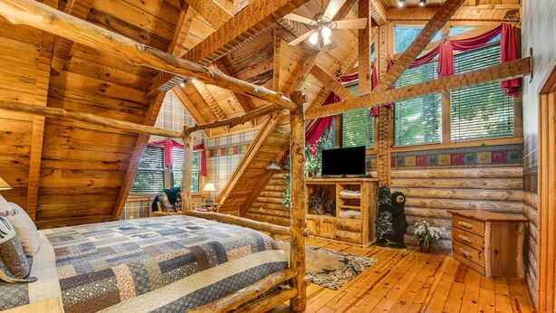 Un dormitorio tipo cabaña de troncos con cama, paredes de madera, ventilador de techo y televisión.