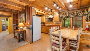 Interior rústico de una cabaña con una mesa de comedor de madera, sillas, un refrigerador y una zona de cocina con armarios de madera y una ventana con persianas.
