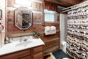 A rustic bathroom with a wooden wall, a white sink, a mirror with a decorative frame, a window with a curtain, and a shower curtain with animal prints. - Sevierville Smoky Mountain Cabin | Hot Tub & Views (Sevierville)