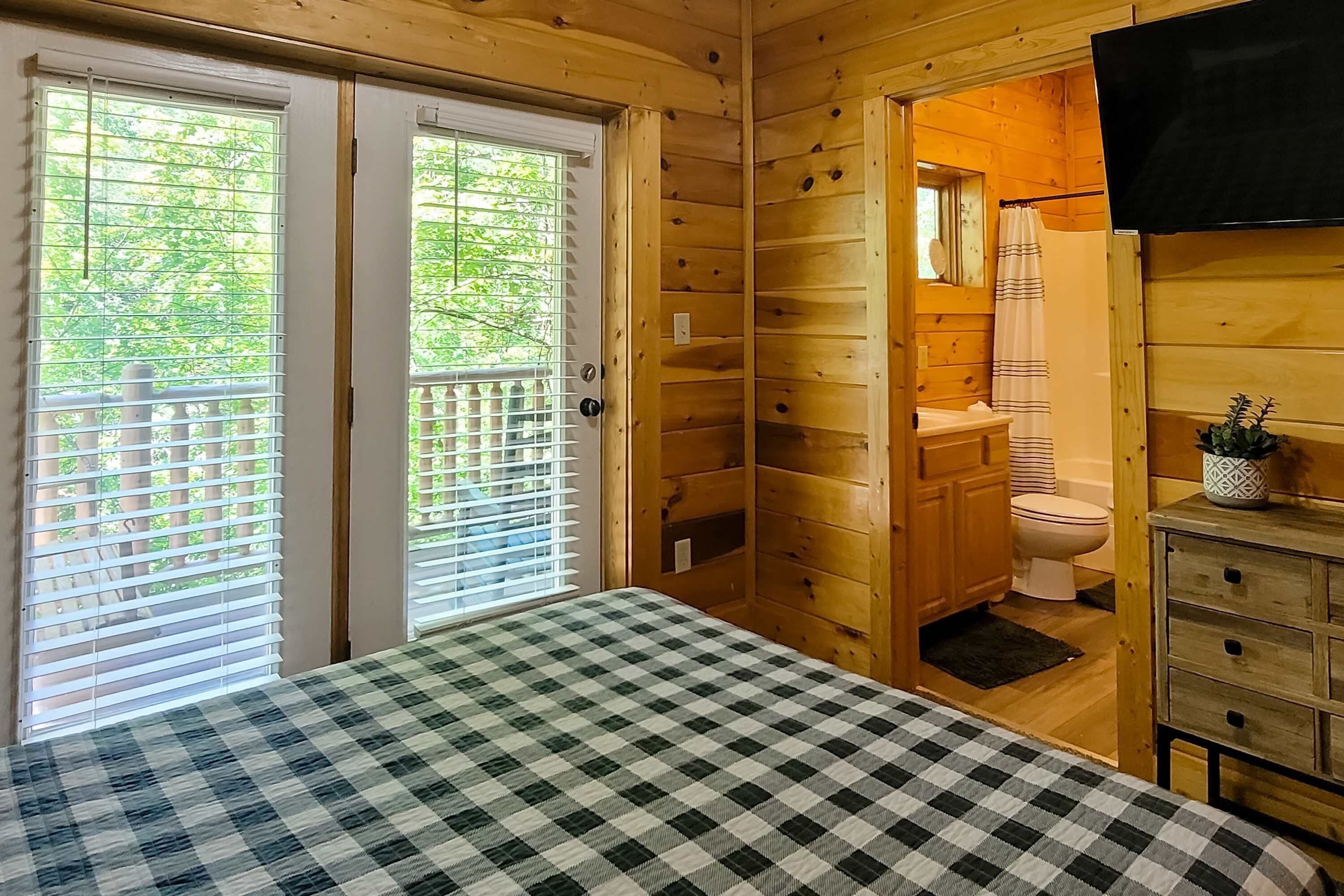 A wooden cabin room with a checkered bed, a wooden dresser, a shower area, and a balcony with a view of trees.