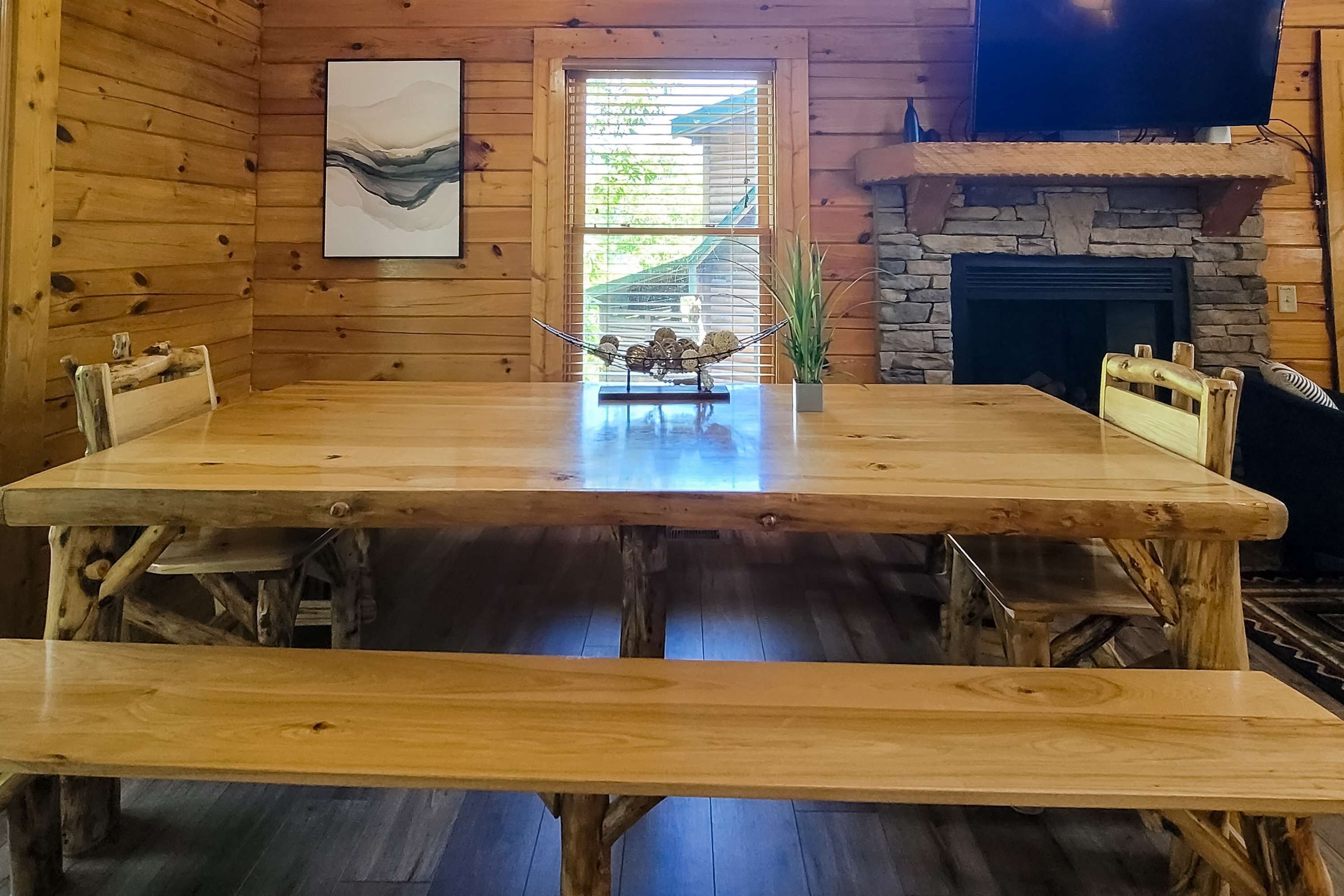 A wooden interior with a large table, benches, a stone fireplace, and a window with blinds.
