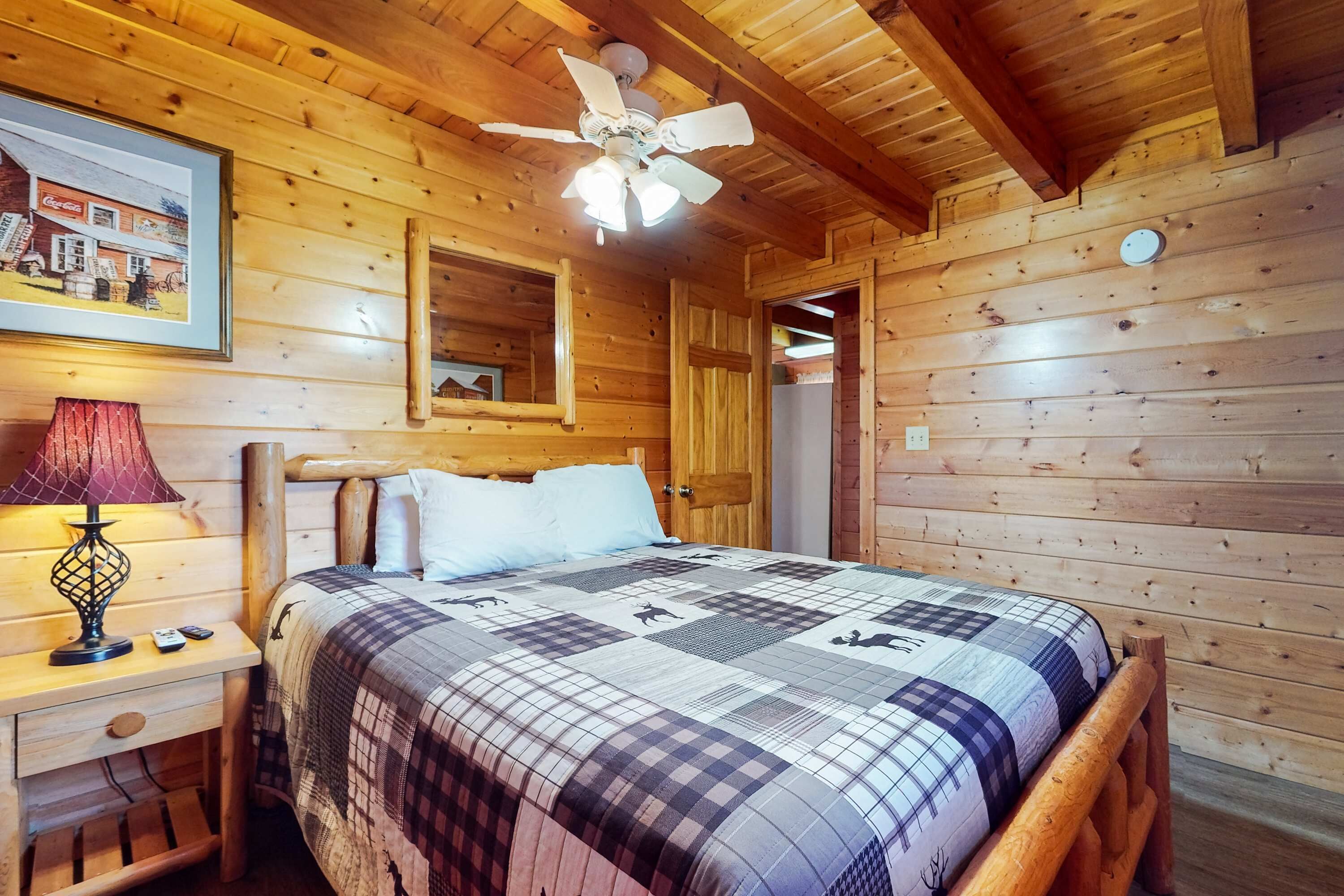 A wooden cabin bedroom with a bed, a ceiling fan, a nightstand, a lamp, and a framed picture on the wall.