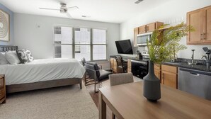 A modern hotel room with a bed, desk, kitchenette, and a large window with blinds. - Landing | Amazing Studio, Gym, Pool (Birmingham)
