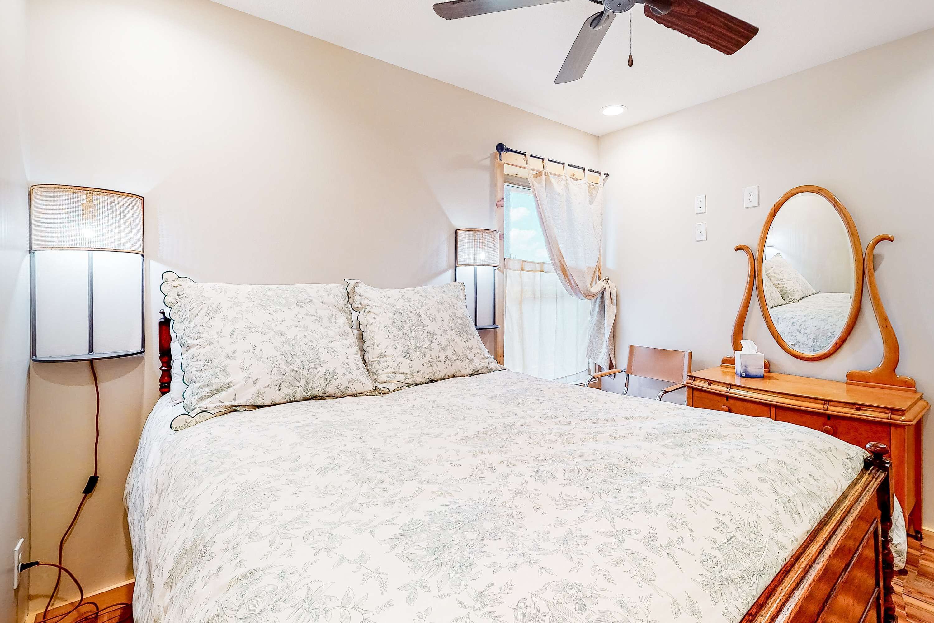 A bedroom with a bed, a wooden dresser, a mirror, a ceiling fan, and a window with curtains.