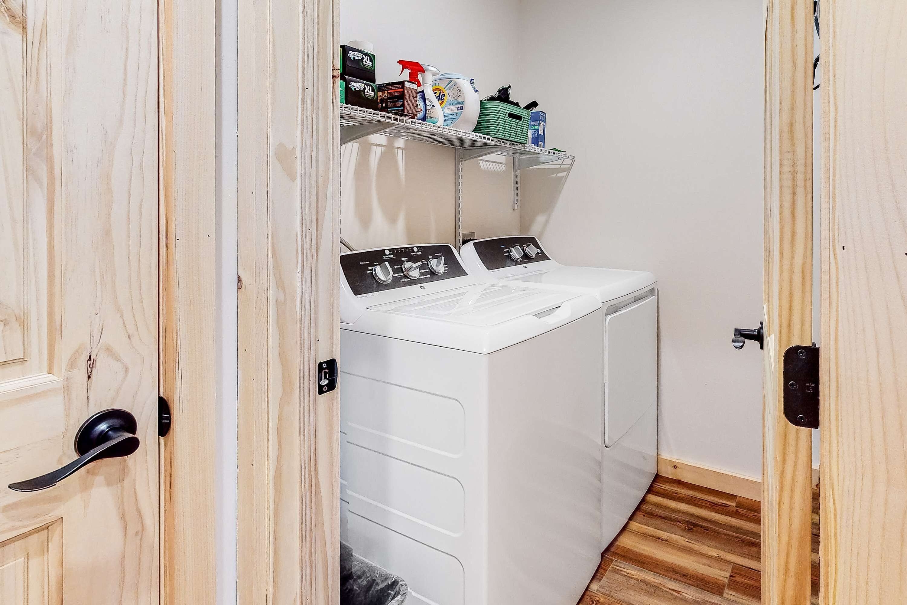 A laundry room with two white front-loading washing machines and a shelf above containing cleaning supplies.
