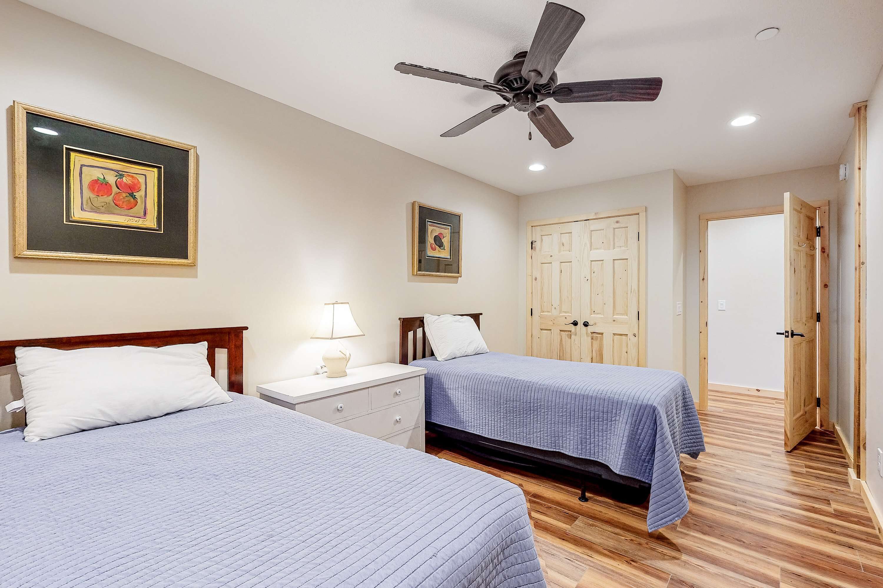 Une chambre avec deux lits, un ventilateur de plafond, un plancher en bois et des œuvres d’art encadrées accrochées aux murs.