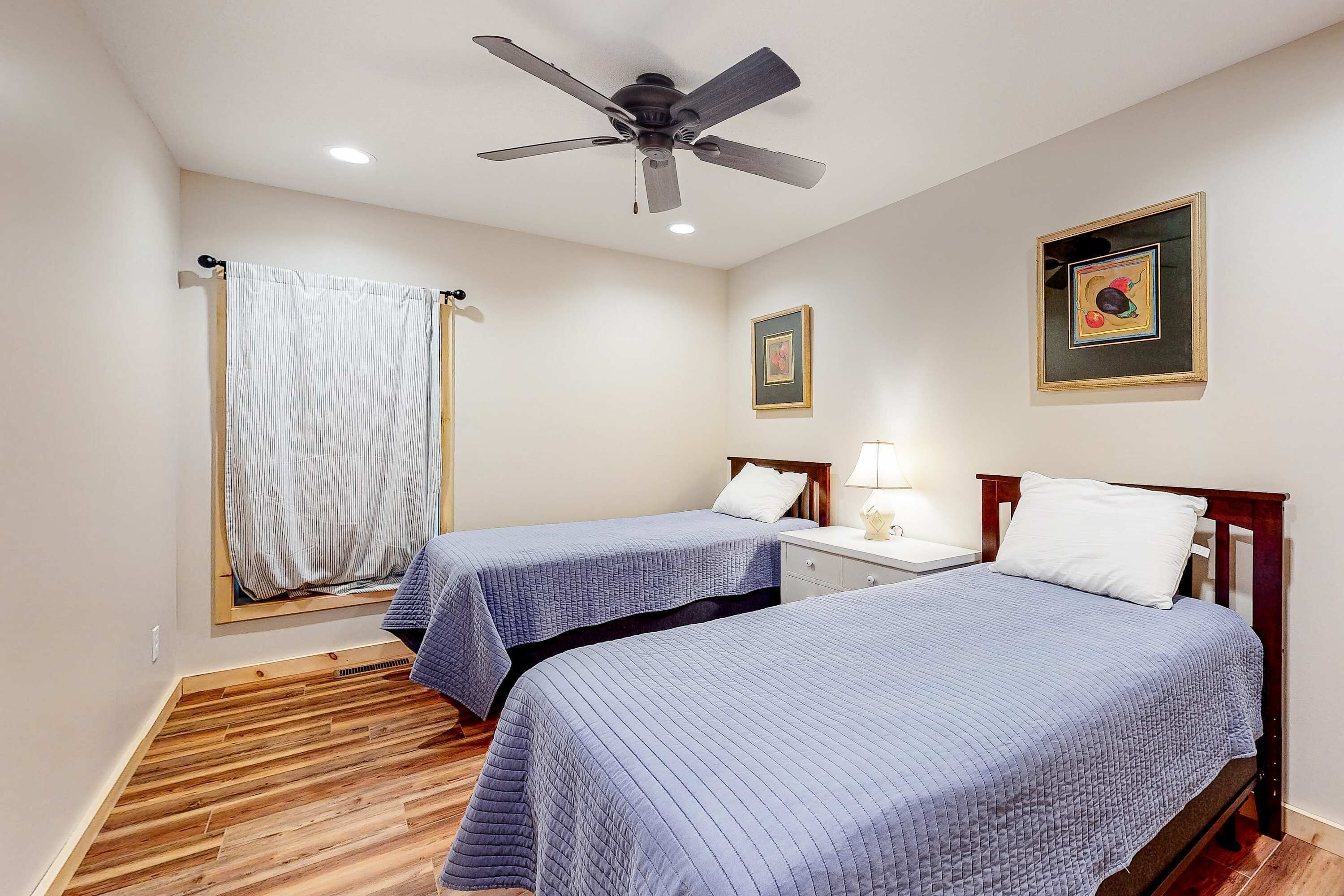 A hotel room with two beds, a ceiling fan, a window with curtains, and a framed picture on the wall.
