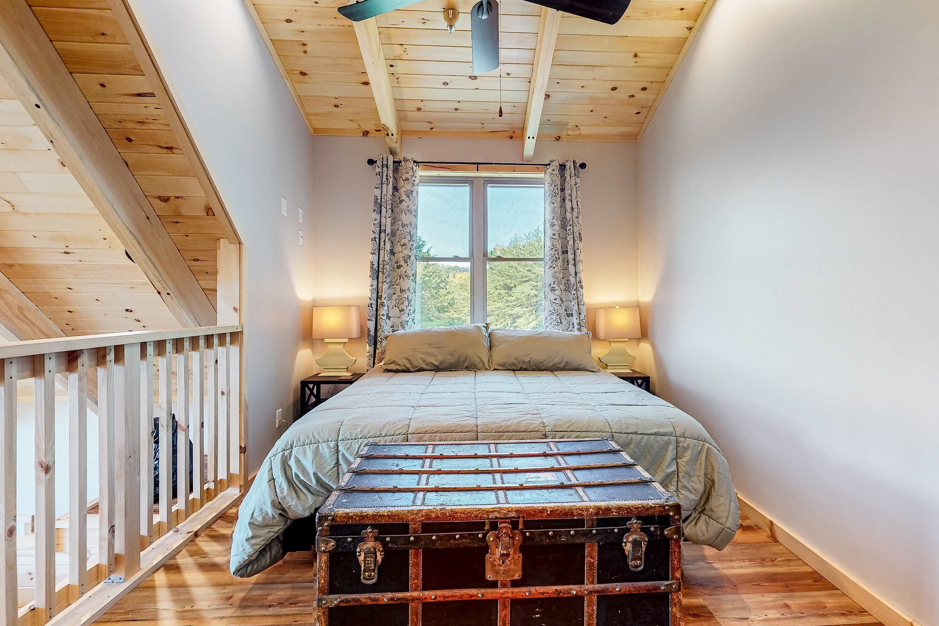 A bedroom with a bed, a window with curtains, a wooden ceiling, and a small table with a lamp.