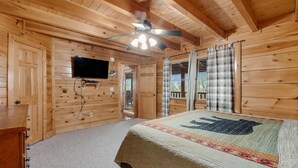 Une chambre dans un chalet en bois, équipée d’un lit, d’un ventilateur de plafond, d’une télévision et d’une fenêtre avec des rideaux.