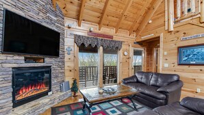 L’intérieur chaleureux d’un chalet avec une cheminée en pierre, un canapé en cuir et un téléviseur fixé au mur.