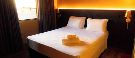 A hotel room with a bed, curtains, a window, and a wall-mounted lamp.