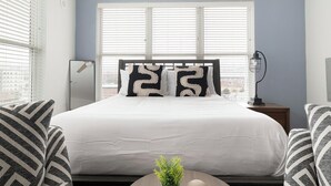 A neatly made bed with white bedding and patterned pillows, a bedside lamp, and a view of the city through large windows. - Landing | Amazing Studio, Pool, Yoga Studio (Birmingham)