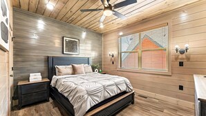 Une chambre avec un plafond en bois, un lit avec une tête de lit, une table de chevet, un luminaire fixé au mur et une fenêtre avec des stores.